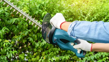 A person wearing gloves skillfully uses an electric hedge trimmer to trim green bushes, showcasing tree trimming expertise in action. The image captures the tool slicing through leaves, highlighting gardening work in progress.